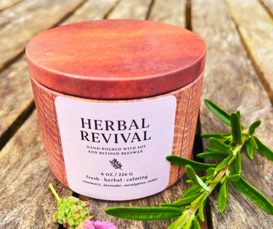 Candle labeled 'Herbal Revival' on a wooden surface with greenery.