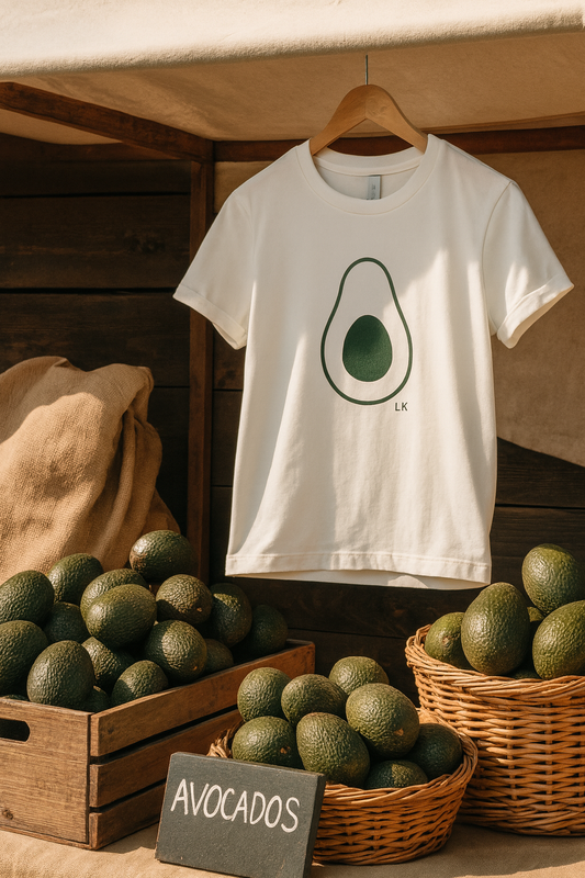 White t-shirt with avocado graphic hanging among avocados on a wooden surface