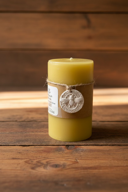 Pure beeswax pillar candle wrapped in a brown kraft paper band with a white circular bee ornament tied around it, sitting on a white textured surface with a blurred background.